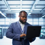 Computer scientist in data center using diagnostic tools and system monitoring software on laptop to identify cause of errors. IT specialist in server farm establishment repairing equipment, camera B